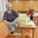 Starejša gospa, oblečena v cvetlično bluzo in črtast predpasnik, uporablja starinsko ročno napravo za rezanje testenin. Njene roke so spretne pri delu, medtem ko v ozadju druge ženske nadaljujejo pripravo testenin na dolgih mizah, pokritih z razvaljanim testom.