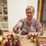 Starejša gospa s kratkimi svetlimi lasmi in očali sedi za leseno mizo ter drži šopek pisanih rož iz krep papirja. Oblečena je v vzorčasto bluzo in se prijazno smehlja. Na mizi so razporejeni številni drugi šopki in prazna pletena košara. V ozadju je bela omara s policami, kjer so postavljeni okrasni predmeti in cvetje.