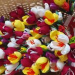 Pletena košara, napolnjena s pisanimi rožami iz krep papirja v rdeči, rumeni, beli in vijolični barvi. Rože so skrbno izdelane, nekatere imajo priložene rdeče kartončke z napisi. Košara stoji na leseni površini, rože so umetelno razporejene.