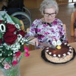 Starejša gospa s sivo pričesko in očali sedi za mizo in reže čokoladno torto z napisano številko 90. Ob njej je šopek rdečih vrtnic v stekleni vazi.
