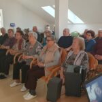 Starejši obiskovalci doma sedijo v tišini in zbrano gledajo filmsko projekcijo. V ospredju je prenosni računalnik, na katerem je viden prizor iz filma. Prostor je svetel, z mansardnimi okni in sodobno notranjo opremo.