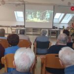 Pogled od zadaj na skupino starejših oseb, ki sedijo pred veliko platno in gledajo film. Na platnu je prizor z otroki v naravi. Prostor je domače urejen, svetel, z dekoracijami in knjižnimi policami v ozadju.