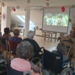 Starejši stanovalci doma, nekateri v invalidskih vozičkih, gledajo film, predvajan na velikem platnu. Prostor je okrašen z ročno izdelanimi dekoracijami in je topel ter prijeten. Na platnu je prizor z dvema fantoma na tleh v naravi.