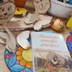 Na leseni površini je razstavljen pisan velikonočni aranžma z ročno izdelanimi zajčki, pisanimi jajčki, papirnatimi metulji in knjigo z naslovom Slovenske narodne pravljice. V ozadju so dodatne dekoracije in okraski.