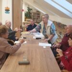 Skupina starejših ljudi sedi za leseno mizo v prijetnem prostoru z veliko naravne svetlobe. Ena izmed žensk stoji in ponuja krožnik s sveže pripravljenimi flancati. Na mizi so kozarci vode, papirji in rolica papirja. V ozadju so police s knjigami, rastline v loncih in stenske dekoracije.