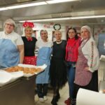 Skupina šestih žensk stoji v profesionalni kuhinji in se nasmehne v kamero. Dve nosita modre predpasnike in bele kuharske uniforme, ena je oblečena kot Mini Miška z rdečo pikčasto pentljo in krilom, druga ima črno krilo iz tila, ena je v rdeče-črni karirasti srajci, zadnja pa v tradicionalni narodni noši s cvetličnim vzorcem. Na pultu pred njimi so pladnji s sveže pečenimi krofi.