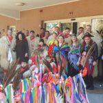 Skupina ljudi, zbranih pred stavbo, verjetno med pustnim ali kostumiranim dogodkom. Nekateri so oblečeni v tradicionalne kurent kostume iz krzna, drugi pa v različne kostume, kot so pirati in cvetlične obleke. Skupina pozira za fotografijo pred pisanimi dekoracijami, kot so trakovi in praznični voziček.