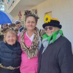 Skupina treh oseb, vključno z oskrbovalko v roza obleki, pozira skupaj na balkonu. Ena oseba je oblečena v kostum z ušesi, druga pa v klobuk, ki spominja na cvet sončnice. V ozadju je videti še druge, ki uživajo v praznovanju.