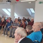 Večja skupina stanovalcev doma pozorno spremlja program. Nekateri sedijo na stolih, drugi v invalidskih vozičkih. Vsi gledajo proti odru, kjer poteka nastop.