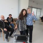 Dve starejši gospe se objemata in nasmejano gledata v kamero. Ena nosi dolgolaso rjavo lasuljo in se opira na hoduljo, druga pa ji prijazno maha v objektiv. V ozadju sedijo še drugi stanovalci, ki se smejejo in uživajo v druženju.