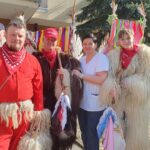 Skupina kurentov v tradicionalnih oblačilih stoji pred domom za starejše. Eden nosi rdečo obleko, drugi so v črnih ali belih kožuhih. Z njimi stoji vodja enote doma za starejše, nasmejana v beli uniformi. V ozadju so trakovi v različnih barvah, smreka in del stavbe.