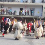 Kurenti v pustnih opravah plešejo in izvajajo svoj obred pred domom za starejše. Na balkonu opazuje več stanovalcev in osebja, nekateri z nasmehi in v pustnih maskah. Okolica je okrašena z barvitimi papirnatimi cvetovi in trakovi.