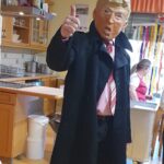 Oseba, oblečena v kostum, ki posnema znanega političnega predstavnika (z masko Donalda Trumpa), stoji v kuhinji. Oseba daje znak z dvignjenim palcem in nosi črni plašč čez srajco in kravato. V ozadju je vidna še ena oseba, verjetno ki pripravlja ali dela v kuhinji.
