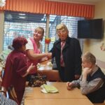Oseba v kostumu Donalda Trumpa se pozdravlja z drugo osebo v prostoru. V ozadju so številni starejši ljudje, nekateri sedijo, drugi stojijo, vse v prostoru, ki je videti okrašen za praznično prireditev.
