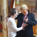 Oseba v kostumu Donalda Trumpa se pogovarja z starejšo žensko v hodniku. Ženska se drži za hoduljo, medtem ko z moškim izmenjujeta prijazen pogovor, moški pa daje znak z dvignjenim palcem. V ozadju so vidne okrasne papirnate verige, ki visijo s stropa.