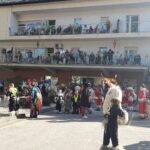 Živahna pustna scena pred stavbo. Ljudje, oblečeni v kurent kostume (krznene obleke), so zbrani pred stavbo, drugi pa v pisanih kostumih, kot so pirati, cvetlični kostumi in drugi ustvarjalni preobleki. Na balkonih nad njimi so baloni in praznične dekoracije, medtem ko ljudje spodaj komunicirajo in se pripravljajo na praznovanje.