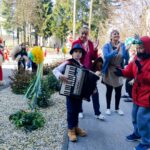 Ljudje, ki sodelujejo v pustnem praznovanju zunaj. Otrok igra akordeon, obkrožen z drugimi osebami, med njimi tudi nekateri v kurent kostumih. Ljudje se zdijo, da uživajo v glasbi in plesu na soncu, z veseljem in prazničnim vzdušjem.