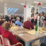 V skupnem prostoru doma starejši stanovalci sedijo za praznično pogrnjenimi mizami, ob katerih nastopa otroški pevski zbor. Mize so okrašene z nageljni, servietami in pijačo, vzdušje je veselo in sproščeno.