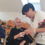 Simpatičen trenutek, ko oskrbovanka doma izroča rožo starejšemu gospodu v invalidskem vozičku. Oba se smehljata, vzdušje je toplo in prijateljsko.
