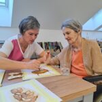 Starejša gospa v bež jopici in oranžni majici sedi za mizo ter se zbrano posveča slikanju. Ob njej je negovalka v roza-beli uniformi, ki jo nežno vodi pri ustvarjanju. Na mizi so slike v postopku nastajanja, kozarec vode in paleta z barvami. V ozadju so police s knjigami in mansardna okna, skozi katera prihaja naravna svetloba.