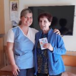 Mlada ženska v svetlo modri in beli uniformi stoji poleg starejše gospe v modrem puloverju. Obe se nasmejano gledata in pozirata za fotografijo. Starejša gospa drži v roki ročno izdelano voščilnico z motivom rož. V ozadju je televizor na steni in koledar.