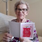 Starejša gospa s kratkimi sivimi lasmi in očali sedi na postelji ter se nasmehne proti kameri. V rokah drži odprto voščilnico s svetlim ozadjem in rdečim motivom vaze z rožami in zvezdami. Oblečena je v bluzo s cvetličnim vzorcem, v ozadju pa je postelja in stenska svetilka.