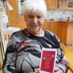 Starejša gospa s kratkimi belimi lasmi sedi na stolu v kuhinji in drži v rokah voščilnico z živahnim rožnatim ozadjem in rjavo vazo s srčki ter rožami. Oblečena je v temno pulover s sivim in rdečim vzorcem. Za njo je kuhinja z lesenimi omarami, pomivalnim koritom in kuhinjskimi pripomočki.
