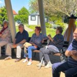 Starejši moški in ženske sedijo na klopci in stolih pod nadstreškom ter opazujejo dogajanje pred njimi. Nekateri ploskajo, ozračje je sproščeno.