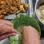 Dve starejši roki mešata maso v zeleni posodi; ob strani lonec z oljem na štedilniku, na pekaču že ocvrti akacijevi cvetovi na papirju za odcejanje.