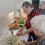 Skupina starejših oseb pripravlja akacijeve cvetove v kuhinji; nekateri mešajo maso, drugi cvetove sušijo ali pripravljajo za cvrtje, na pultu velika skleda cvetov in pladenj s cvrtjem.