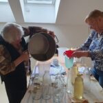 Dve starejši ženski v kuhinji prelivata bezgov sirup iz velike posode v merilni vrč, pripravljeni za polnjenje steklenic.