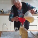 Starejša ženska previdno preliva bezgov sirup iz merilnega vrča skozi lijak v steklenico, na mizi so pokrovčki in prazne steklenice.