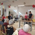 Skupina stanovalcev doma starejših oseb sedi v krogu in posluša predstavitev mladega čebelarja. Na mizi pred njim so čebelarski pripomočki, satnice in kozarci z medom. Ob njem stoji zaposlena v domu, prostor pa je svetel in okrašen z visečimi okraski.
