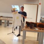 Mladi moški drži v rokah čebelarsko zaščitno obleko s čelado in mrežo. Ob njem je miza s čebelarskimi pripomočki, vključno s satnicami, orodji, kadilnikom in kozarci medu. V ozadju sedijo starejši udeleženci delavnice.