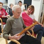 Starejši moški v ospredju sedi in pregleduje čebelji sat, medtem ko ga spremlja medicinska sestra. Okoli njega sedijo druge starejše osebe, nekatere držijo liste z besedilom. Vsi so pozorni na predmet v njegovih rokah.