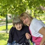Dve ženski, ena uporabnica doma starejših in ena zaposlena, se objemata pod zelenim drevesom na vrtu doma.