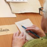 Starejša ženska piše ime Antonija s svinčnikom na črtni papir poleg primerka gotičnega napisa.