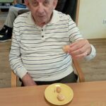 Starejši moški v črtasti majici sedi za mizo, v roki drži miško, pred njim na rumenem krožniku ležijo tri miške. Na mizi je tudi kozarec soka.