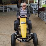 Starejši moški z nasmehom sedi na rumeno-črnem pedalnem gokartu pod nadstreškom doma. Z rokami drži volan in pozira za fotografijo.