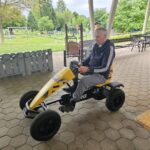 Moški v športni jopici sedi na rumeno-črnem pedalnem gokartu, pripravljen na vožnjo po tlakovani površini. V ozadju so klopi in zelen park.