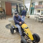 Moški s kapo vozi rumeno-črni pedalni gokart po dvorišču doma, mimo kamnitega vodnjaka. V ozadju drug moški hodi proti vhodu stavbe.