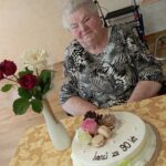 Starejša gospa sedi za mizo, okrašeno z rumeno prtom. Pred njo je rojstnodnevna torta z napisom »Ivanki za 90 let«, okrašena z rožami in makroni. Ob torti stoji bela vaza z rdečimi in belo rožo.