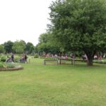 Širši pogled na urejen park z veliko zelenja, kjer se številne osebe, večinoma v invalidskih vozičkih, sprehajajo ob spremstvu. Ozadje prikazuje mirno okolje z drevesi in klopmi.