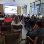 Občinstvo, sestavljeno predvsem iz starejših oseb, sedi v dvorani in spremlja predstavitev filma; dva govorca stojita pred platnom, na katerem je naslov dokumentarnega filma.