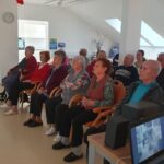Stanovalci doma za starejše sedijo v dveh vrstah stolov in zbrano spremljajo projekcijo filma. Na desni strani je viden računalnik in zvočniki, ki podpirajo predvajanje.