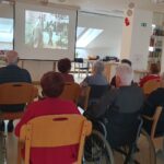 Pogled s hrbtne strani na skupino starejših oseb, ki gledajo film, projiciran na platno. Prostor je svetel in opremljen z domačimi elementi, kot so knjižne police in okrasne luči.