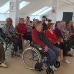 Širši pogled na udeležence kinopredstave, ki sedijo v krogu, večina pozorno spremlja film. Nekateri stanovalci so v invalidskih vozičkih, vzdušje je sproščeno in domače.
