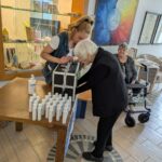 Mlada ženska in starejša gospa si skupaj ogledujeta notranjost UV-škatle na mizi z razkužili, v ozadju še dve stanovalki, ena od njiju na hodulji; dogajanje poteka ob vitrini z ročnimi izdelki.
