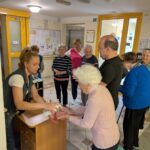 Mlada ženska razkužuje roke starejši gospe, medtem ko drugi stanovalci čakajo v vrsti; prizor poteka na hodniku doma za starejše.