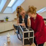 Mlada ženska pomaga starejši gospe pri uporabi črne UV-škatle na mizi, obe z nasmehom gledata v notranjost naprave; svetel prostor z okenskimi strešnimi svetlobami.
